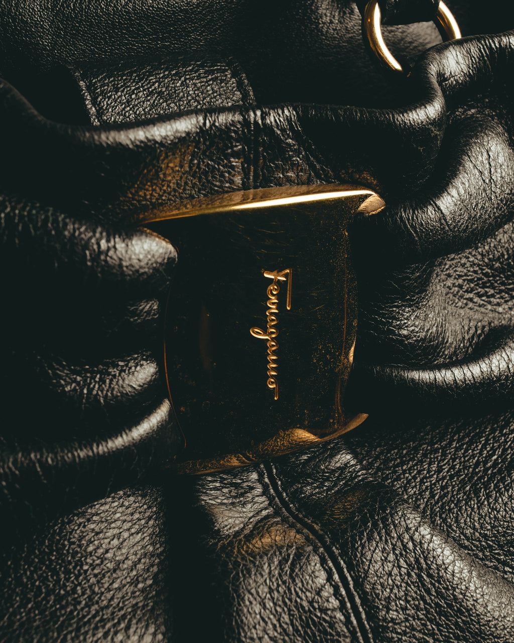 ブラックのギャザーを寄せたレザーボディ中央に配された、筆記体の Ferragamo ロゴ入りゴールドトーンの大型留め具（クラスプ）。周囲には細やかなシボと深いギャザーの陰影が写ります。