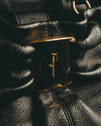ブラックのギャザーを寄せたレザーボディ中央に配された、筆記体の Ferragamo ロゴ入りゴールドトーンの大型留め具（クラスプ）。周囲には細やかなシボと深いギャザーの陰影が写ります。