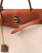 HERMÈS PARISの刻印が入るゴールドカラーのセリエ金具を配した、フロントのベルト式留め具のディテール。赤みを含む深いブラウンのレザートップパーツと、生成りからライトベージュのキャンバス調織地、縦に垂れるクロシェットを確認できます。