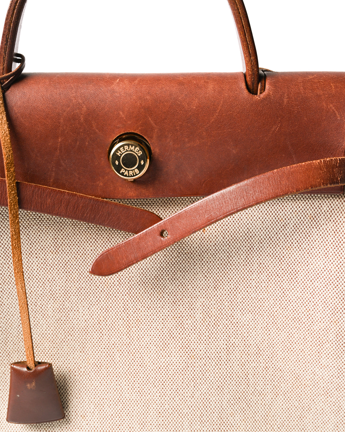 HERMÈS PARISの刻印が入るゴールドカラーのセリエ金具を配した、フロントのベルト式留め具のディテール。赤みを含む深いブラウンのレザートップパーツと、生成りからライトベージュのキャンバス調織地、縦に垂れるクロシェットを確認できます。