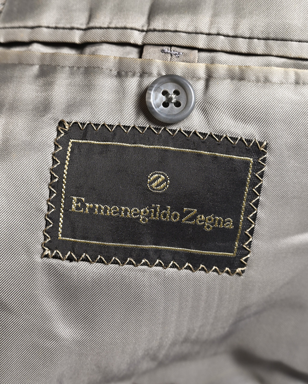 滑らかなグレー系の総裏地に配された、円形のZ意匠を持つErmenegildo Zegna表記の服地ネーム。内ポケット上部の4つ穴ボタンと、ネーム周囲のジグザグステッチも確認できるカット。