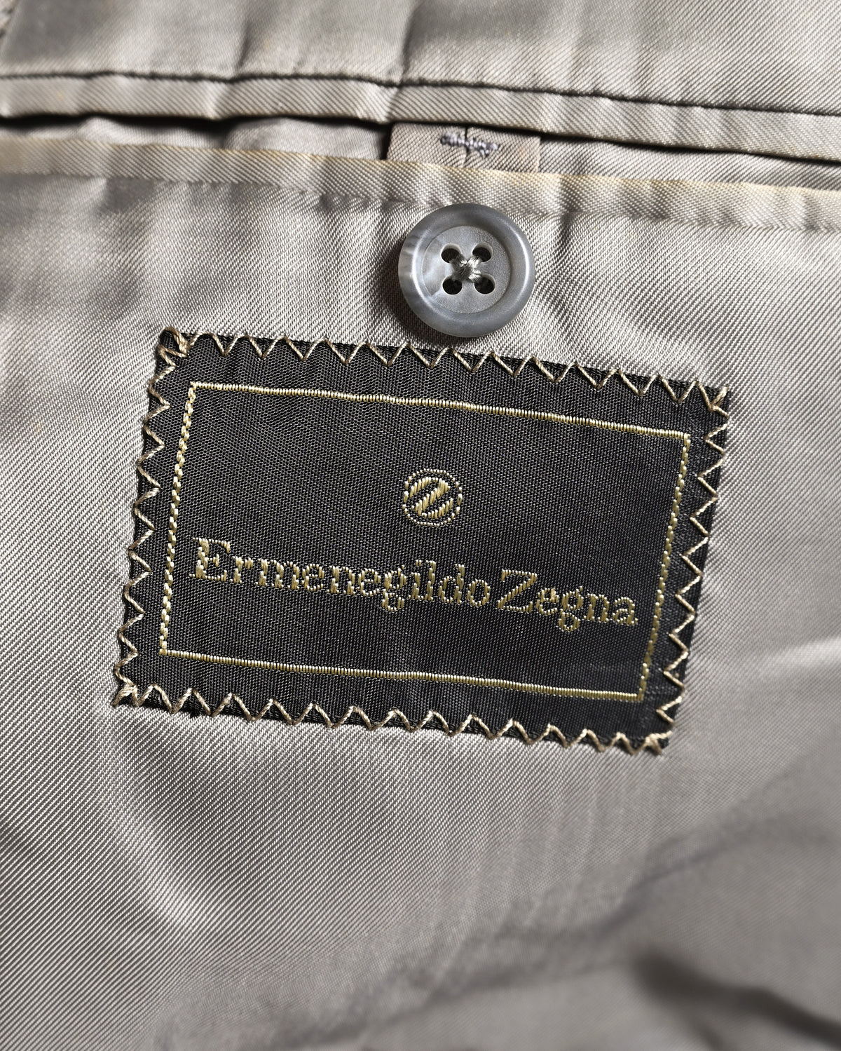 滑らかなグレー系の総裏地に配された、円形のZ意匠を持つErmenegildo Zegna表記の服地ネーム。内ポケット上部の4つ穴ボタンと、ネーム周囲のジグザグステッチも確認できるカット。