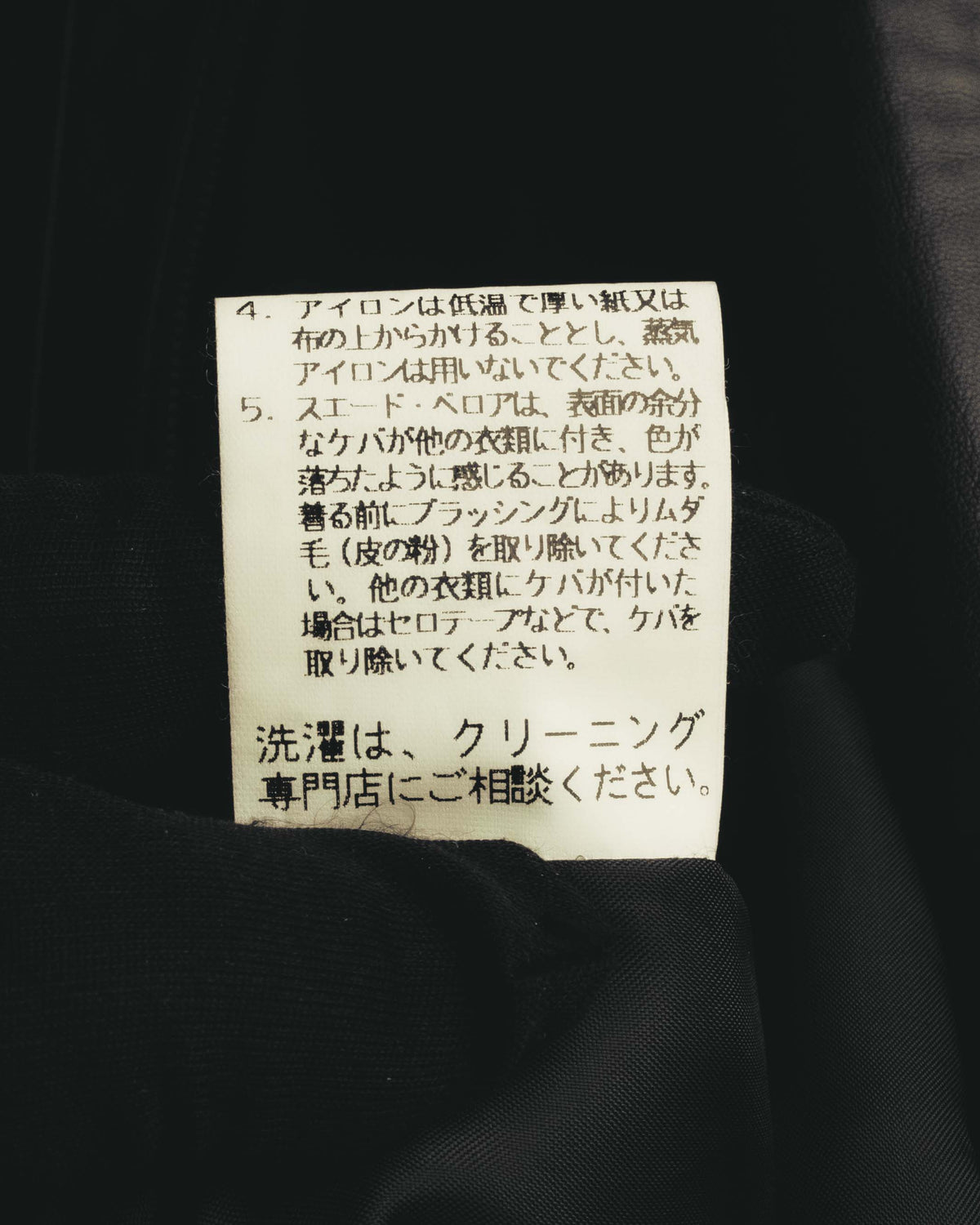 内側に配された洗濯表示タグのクローズアップ。スエード・ベロア表面のケアとクリーニング専門店への相談に関する表記が確認できます。