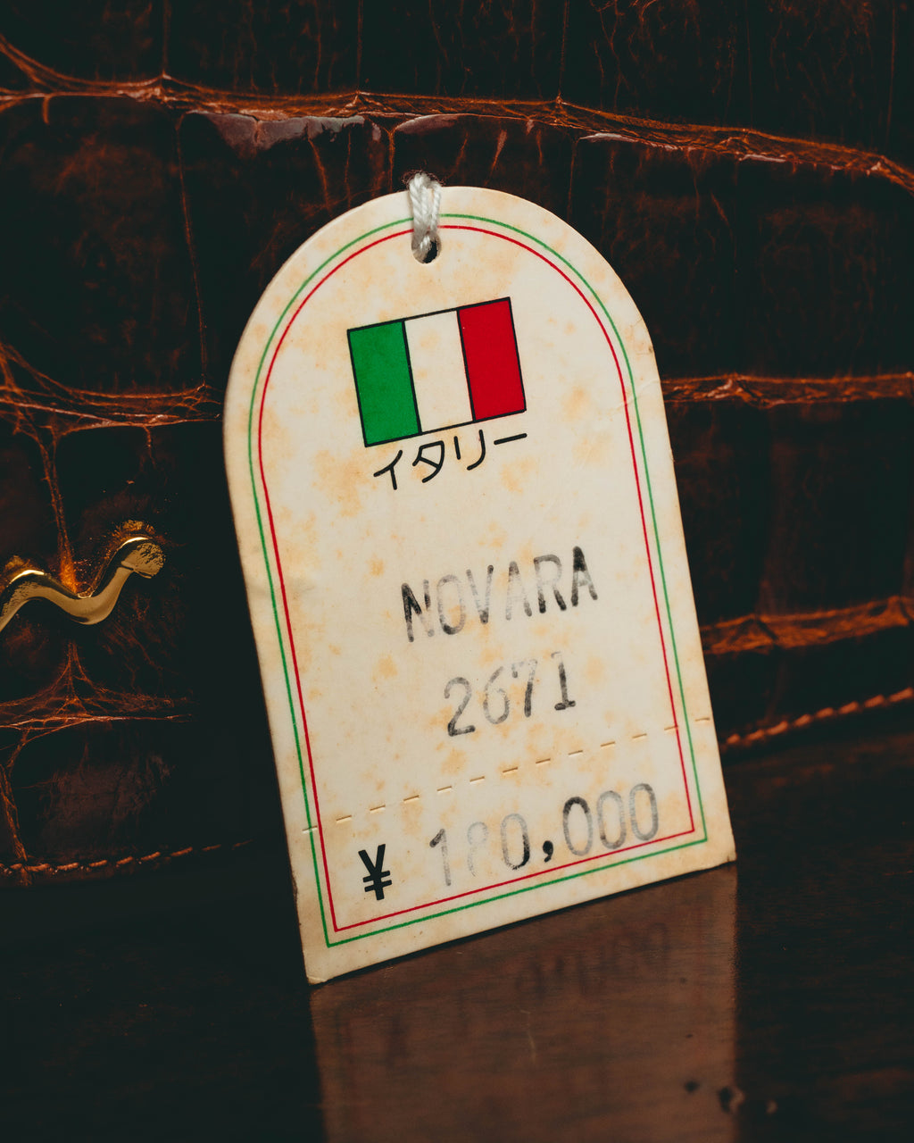 イタリア国旗と「イタリー」、品番「NOVARA 2011」、価格表記が確認できる旧式の紙札。背面にはマホガニーブラウンの鱗状の外装とゴールドカラーのチェーンの一部が写る。