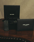SAINT LAURENT PARISのロゴが入ったボックスと保存袋、下げ札を配した付属品のカット。ボックス内にはブラックレザーストラップとYSLモノグラムのメタルパーツが見える。